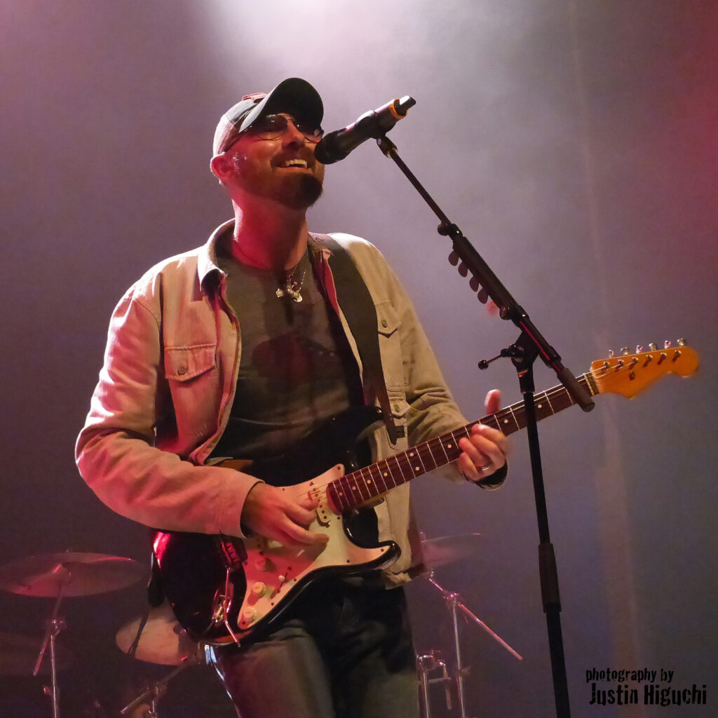 Corey Smith playing a guitar and singing into a microphone in a foggy and dark room.
