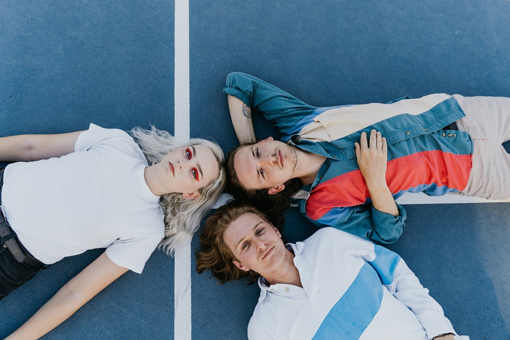 Three members of the band are laying on a tennis court, it is one female and two males, the colors are blue and red