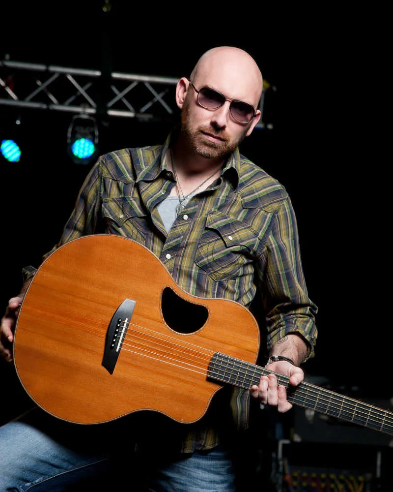 Corey Smith on the stage with a wooden acoustic guitar,he is wearing sunglasses with a green and yellow plaid shirt with jeans.