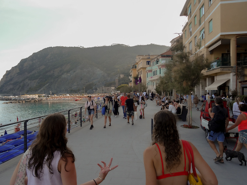 Two girls walking and talking along the buildings that line the water, with a mountian in the background