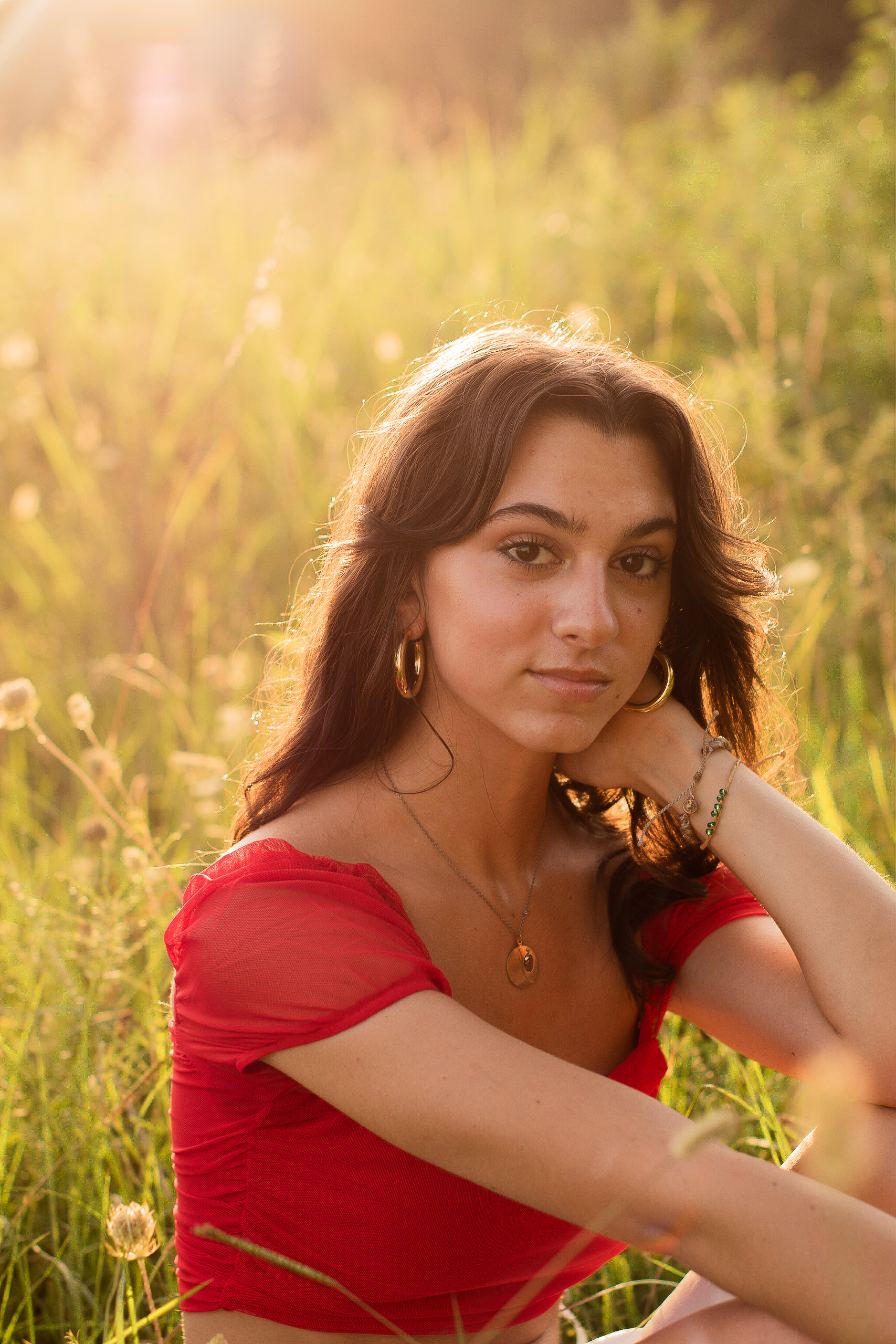 a girl in a red top and white skirt sitting in a sunny feild and looking at the camera