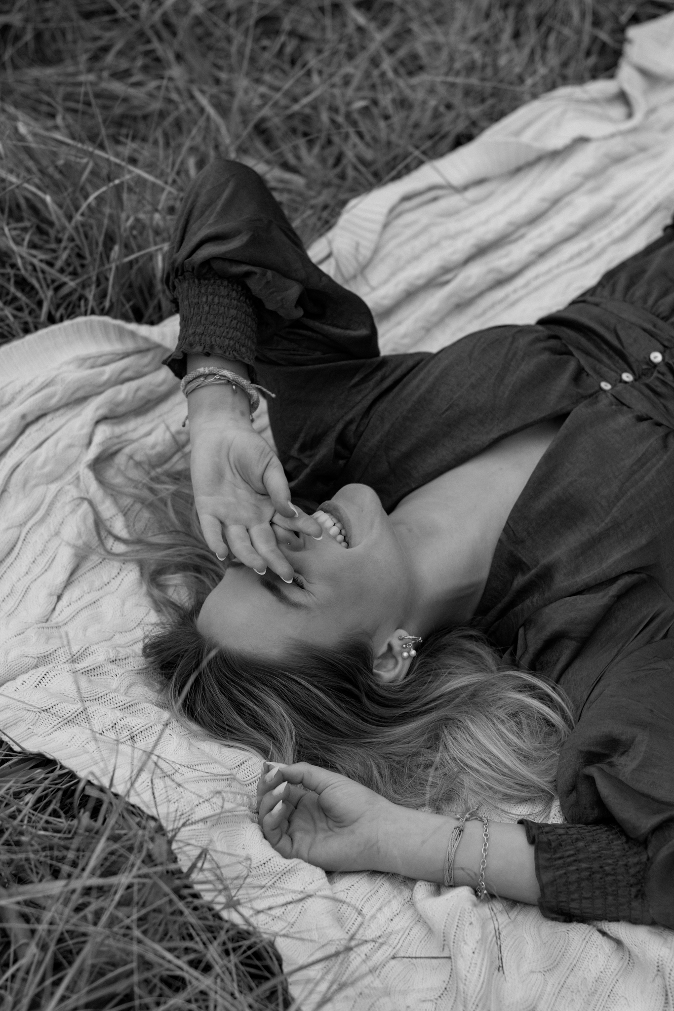 a black and white photo of a girl laying in a feild on a blanket, she is smiling and covering her eyes with her hand