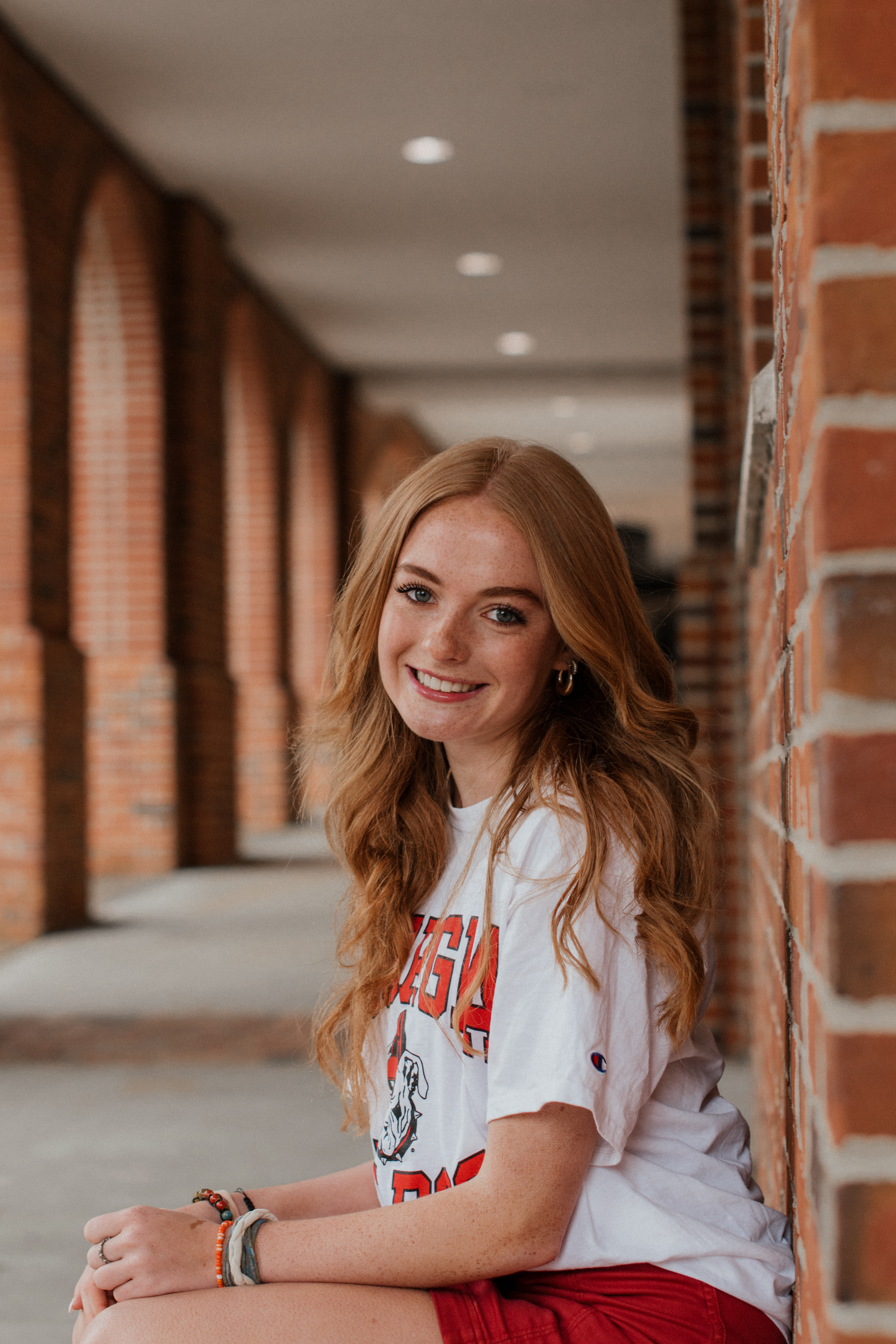 a girl in a UGA shirt leaned up against a brick hallway looking at the camera and smiling