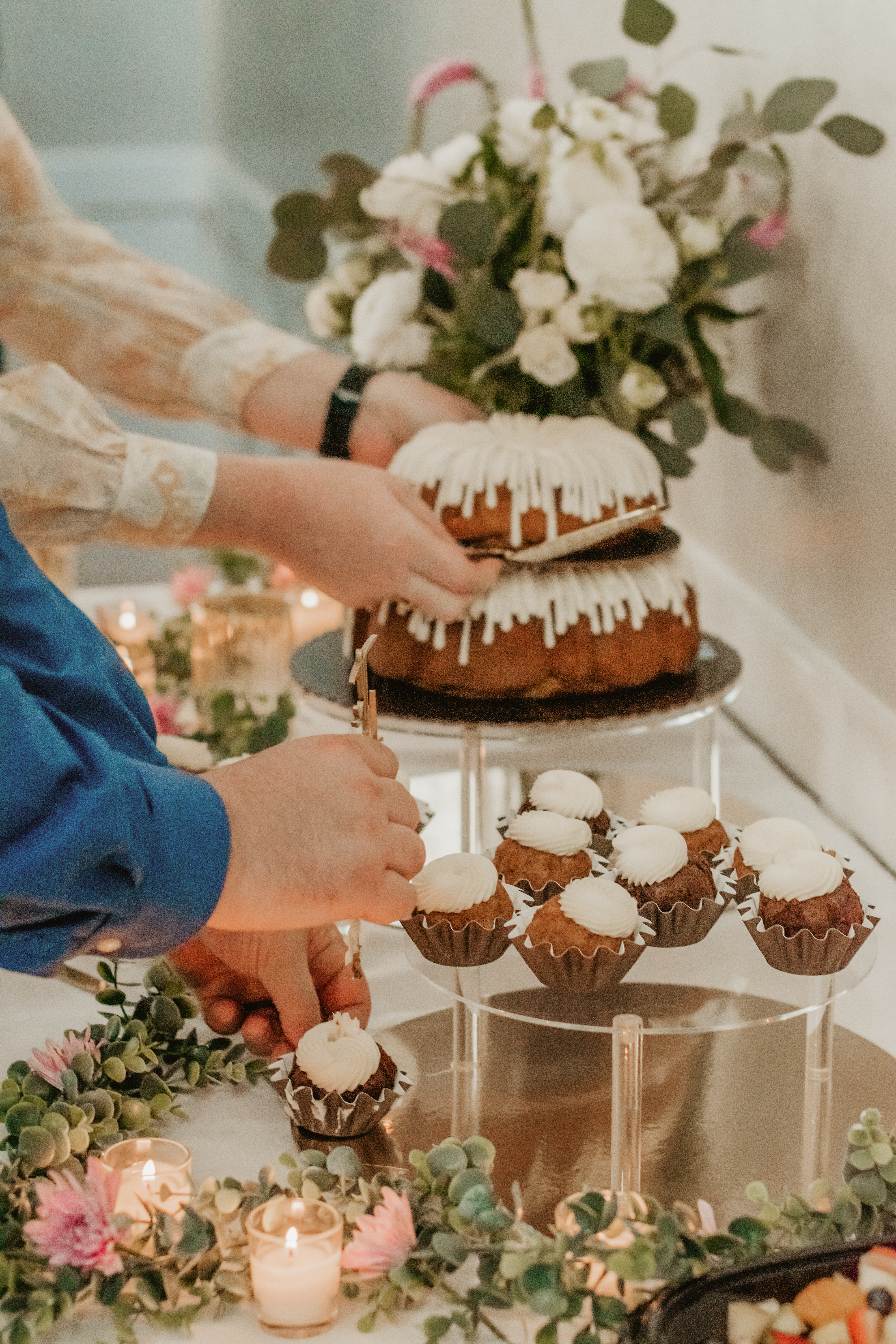 a photo of a wedding bundt cake with cupcakes sitting on a table at the reception, two people's arms are serving the cake