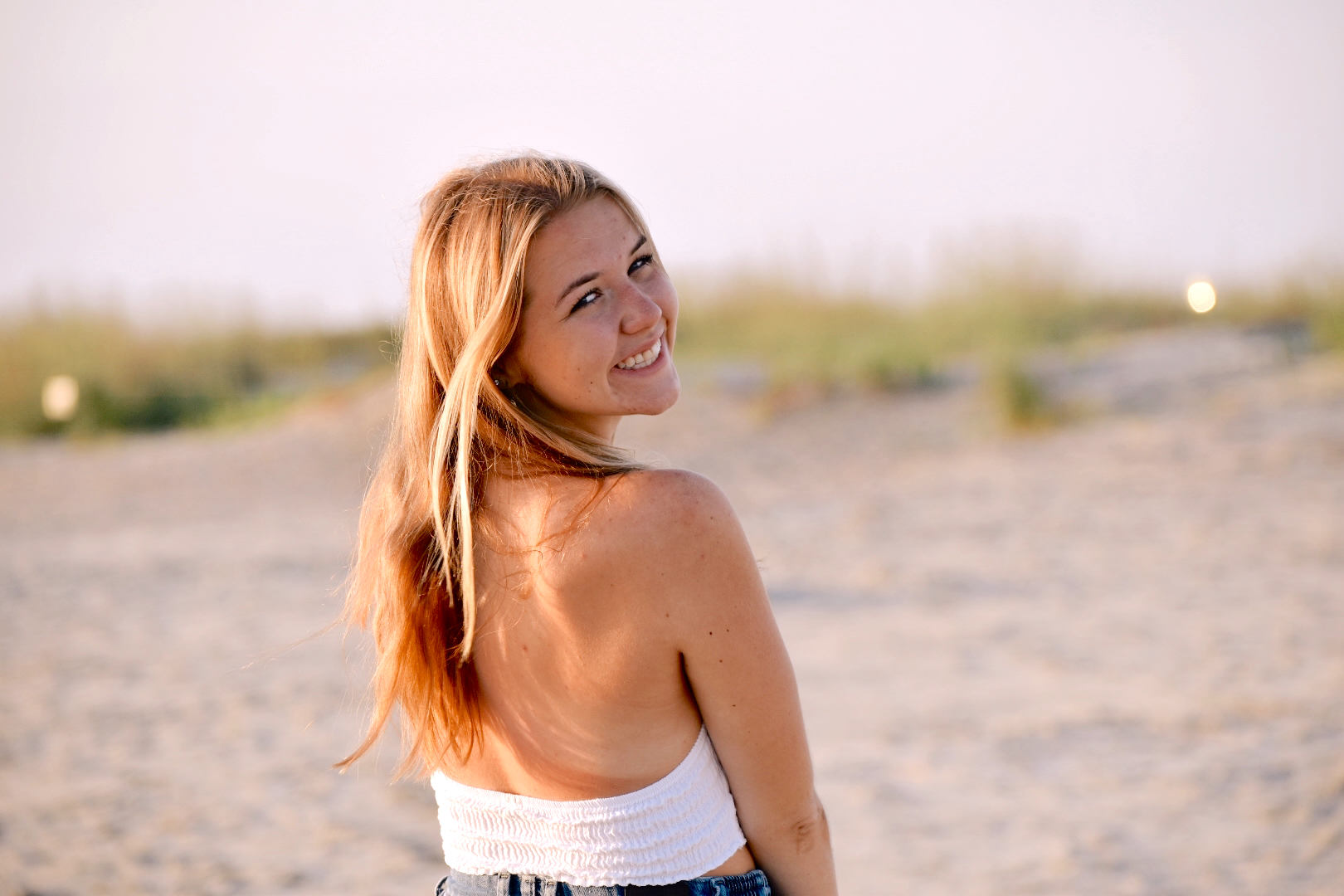 a girl in a white top on the beach, looking over her shoulder and smiling at the camera